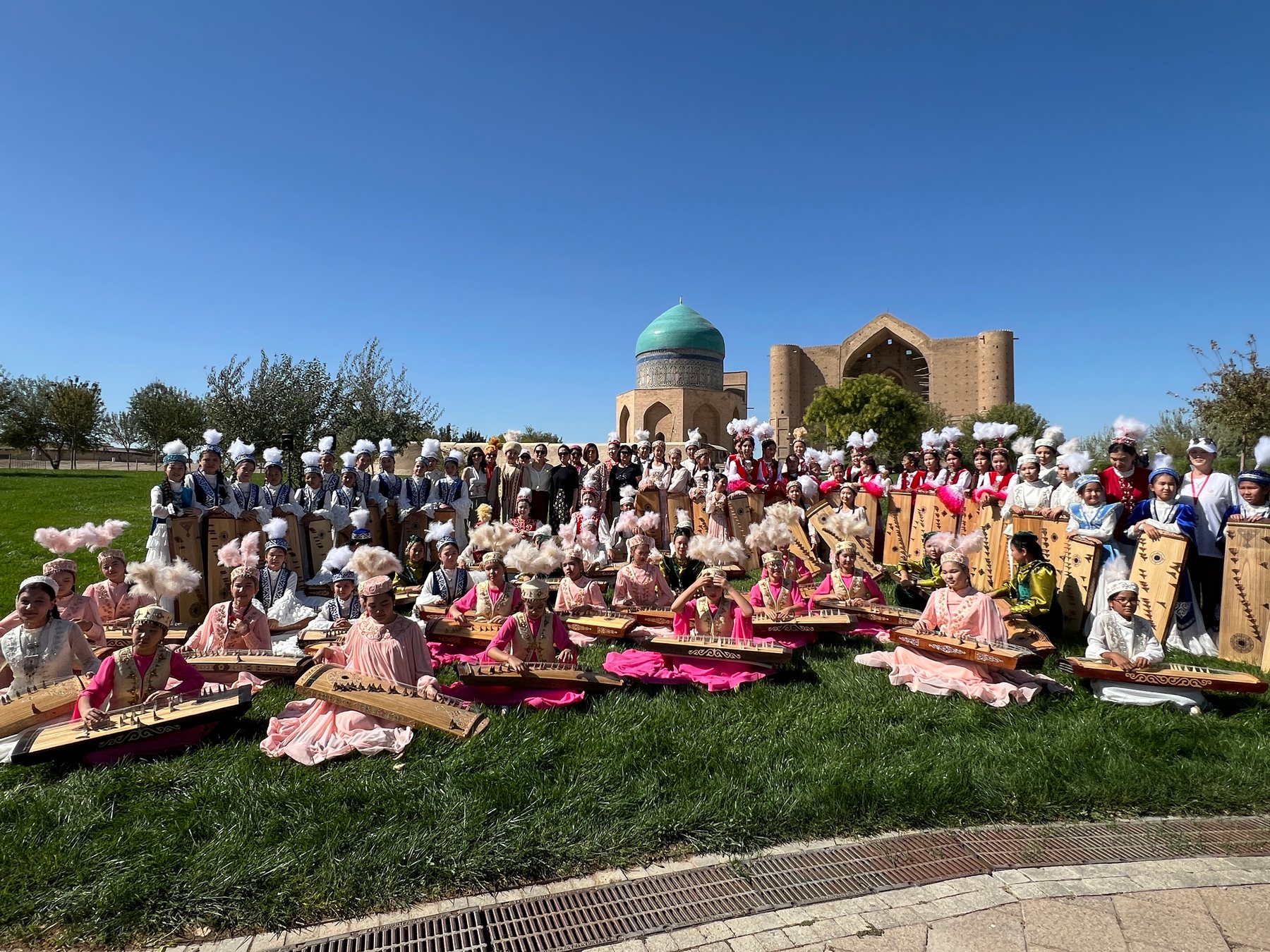 A large group of performers and participants gathered outdoors before historic brick architecture in Kazakhstan.