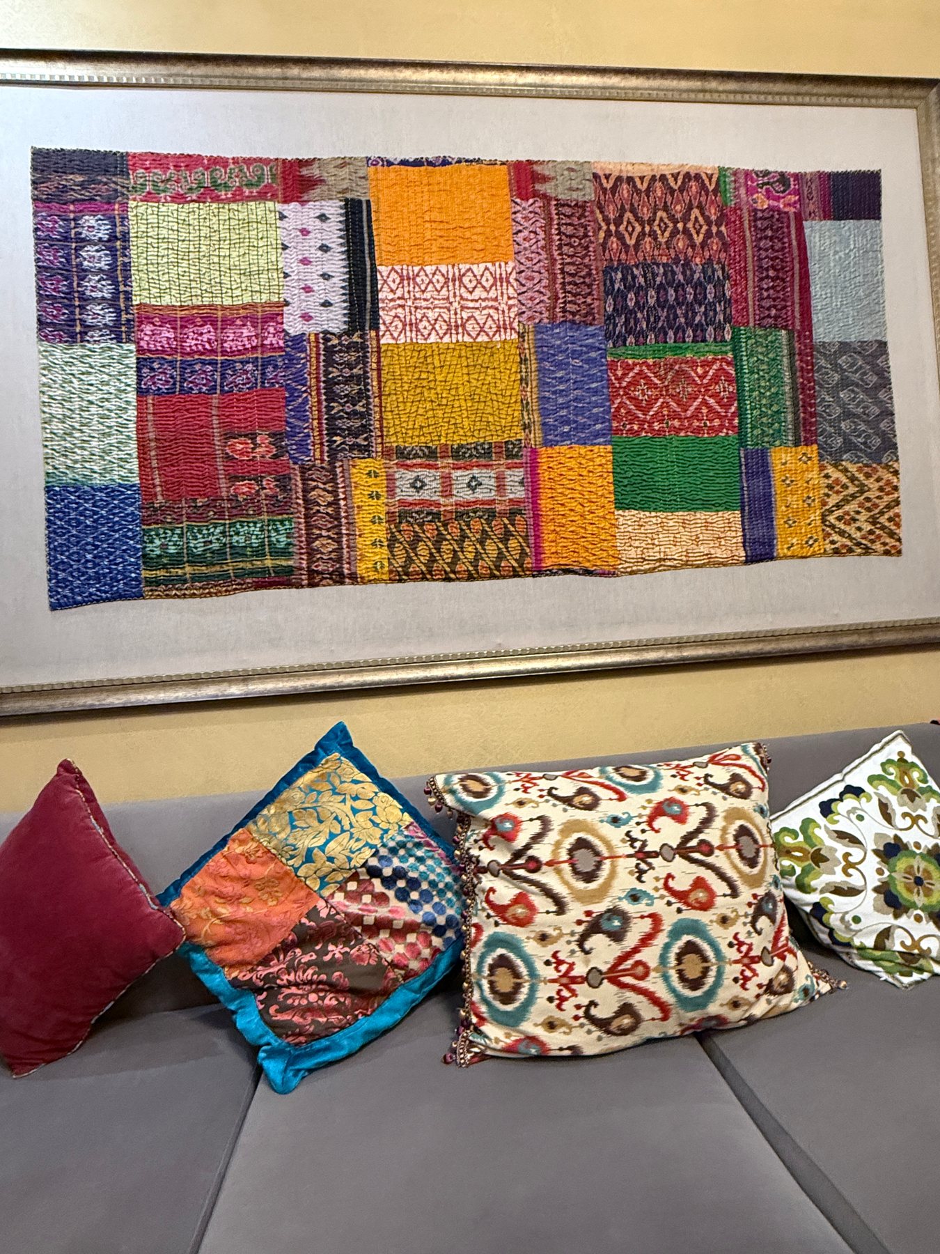 A framed patchwork textile artwork displayed above richly patterned cushions.