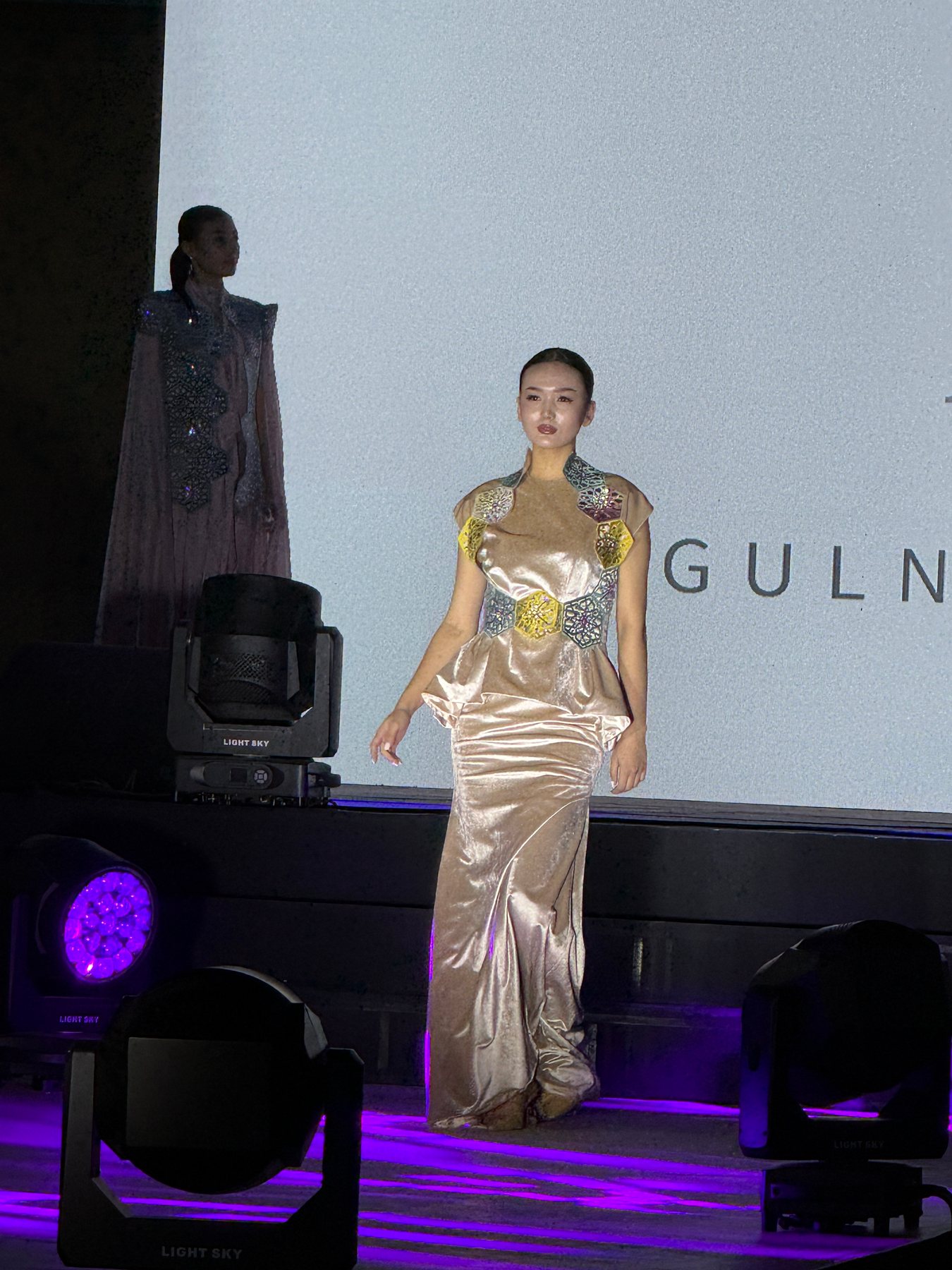 A model presenting a metallic pale-gold gown on an outdoor runway.