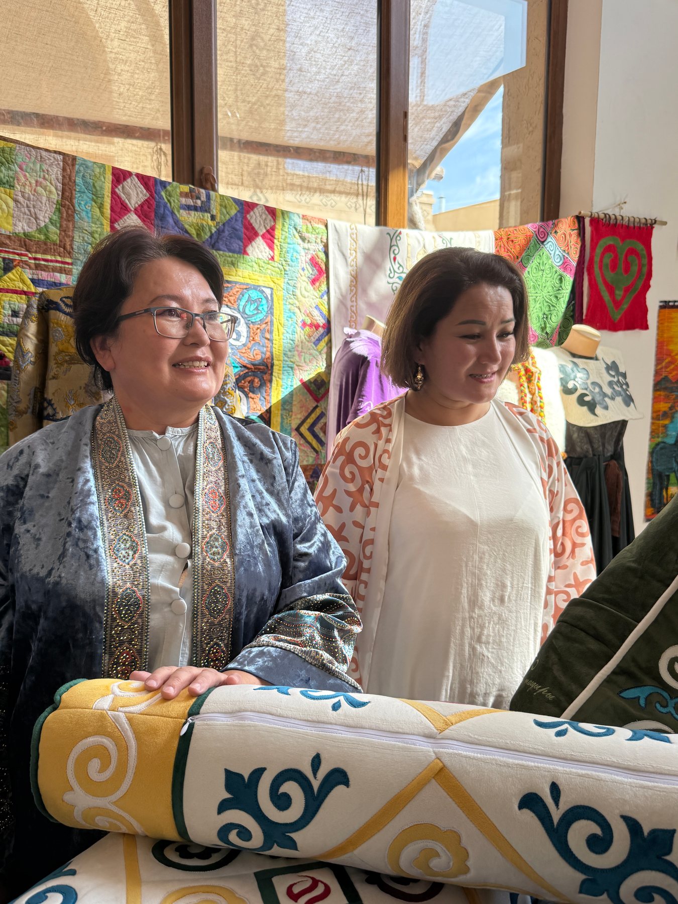 Two women at a textile exhibition with patterned quilts and cushions behind them.