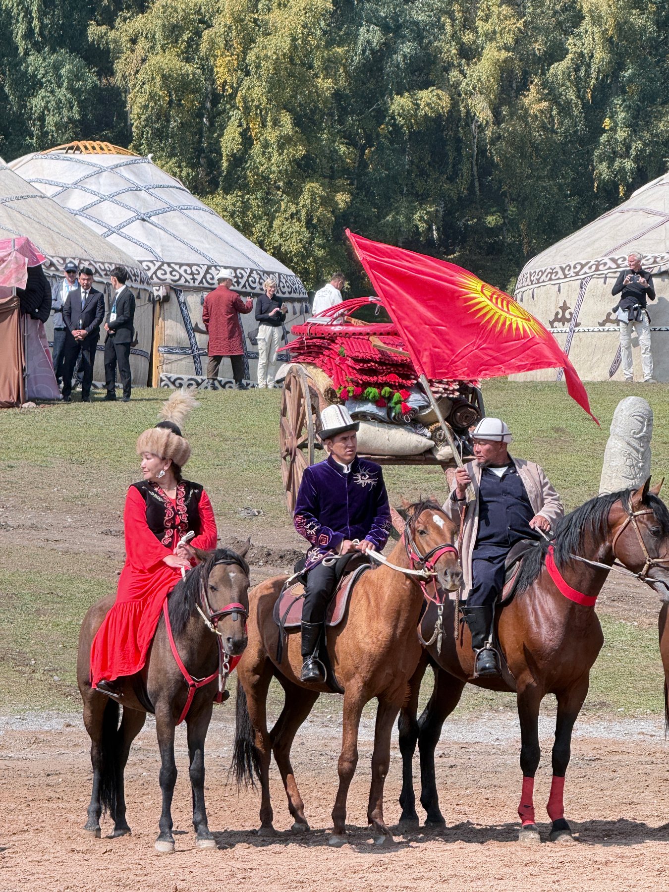 Three mounted participants crossing festival grounds with a large red flag and yurts behind them.