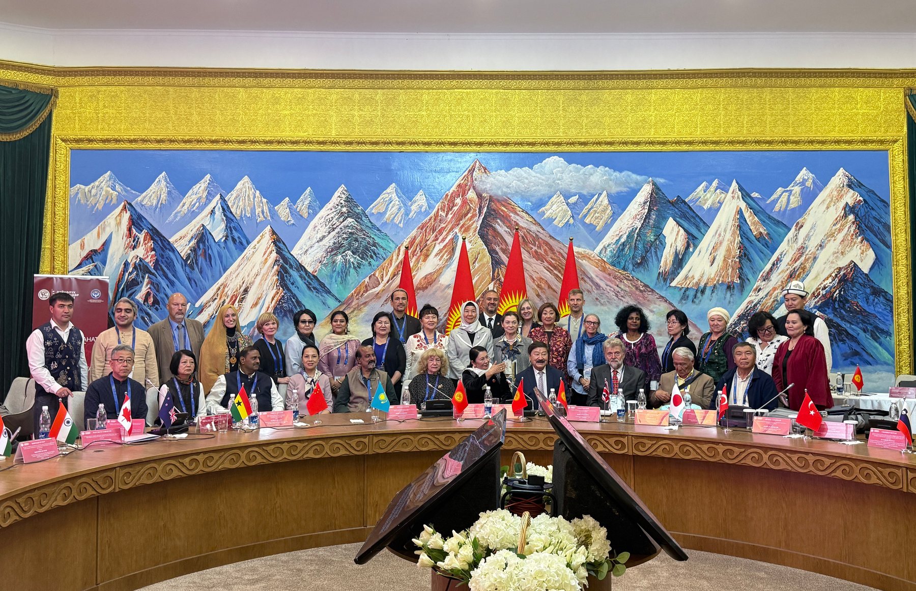 International participants gathered around a conference table before a mountain mural.