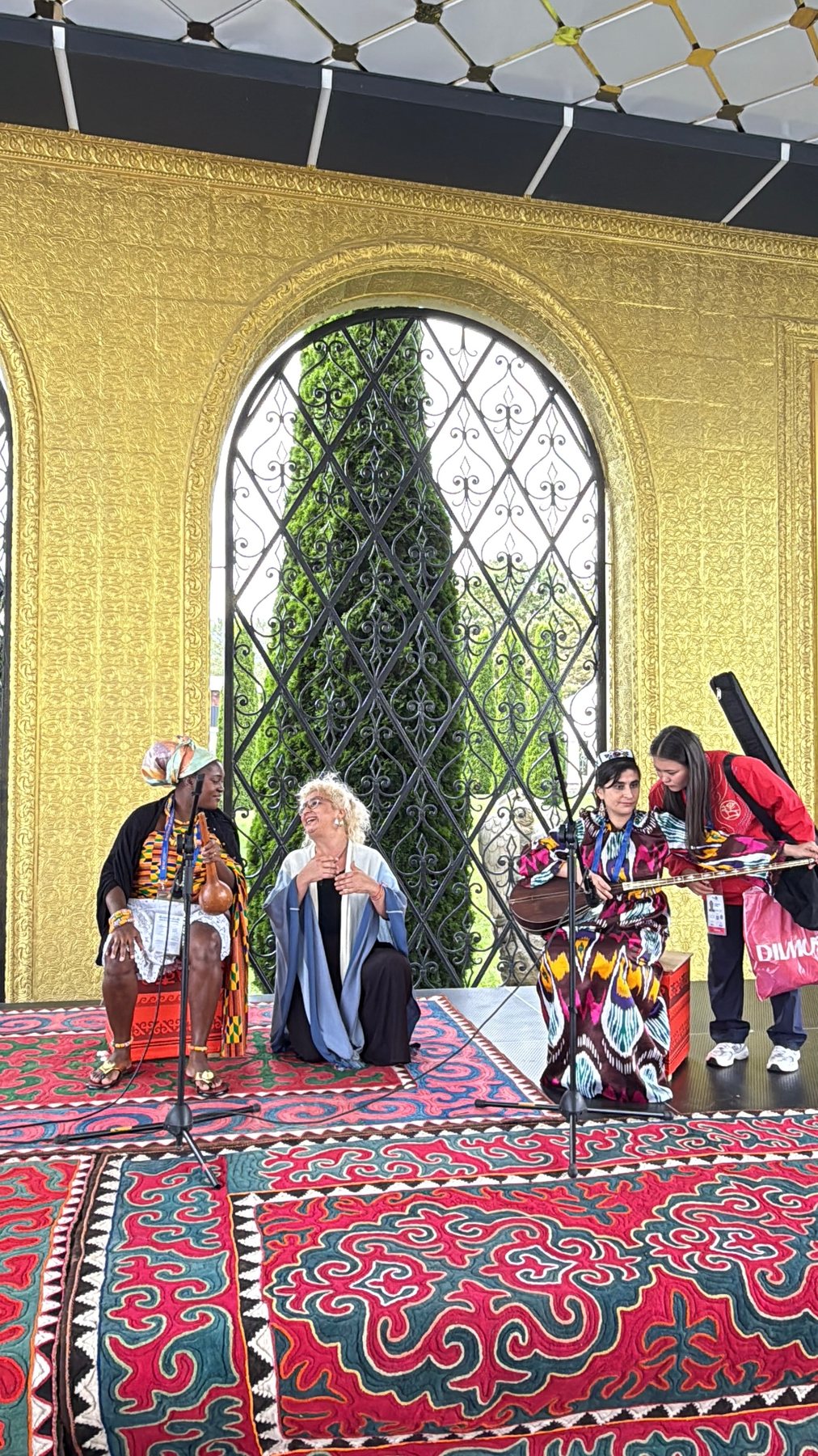 Three international women musicians seated with their instruments among patterned textiles in an ornate pavilion.