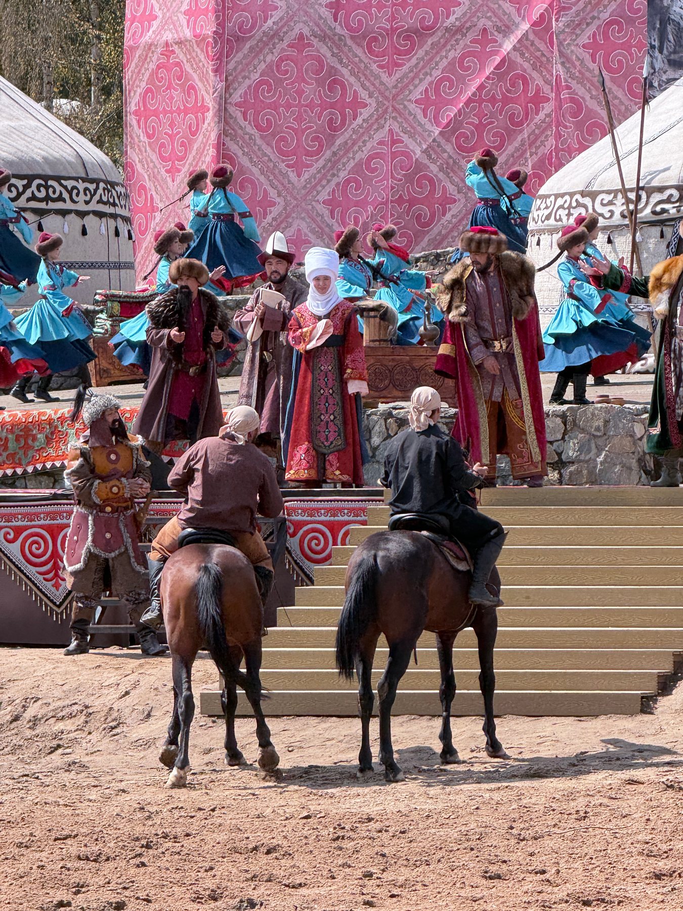 A large staged scene with riders, dancers, yurts and a patterned mountain backdrop.