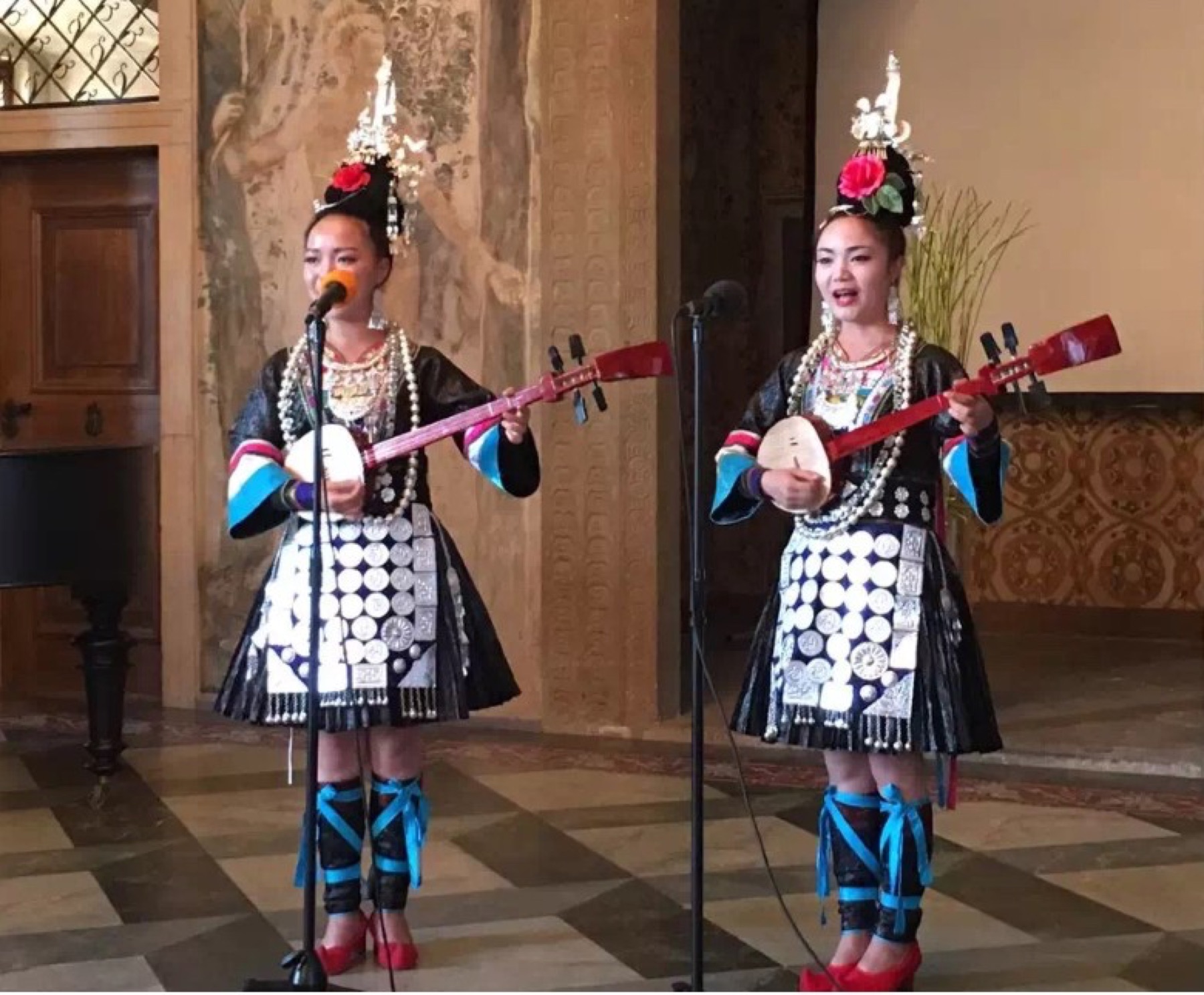 Two musicians in decorated performance dress sing and play long-necked string instruments.