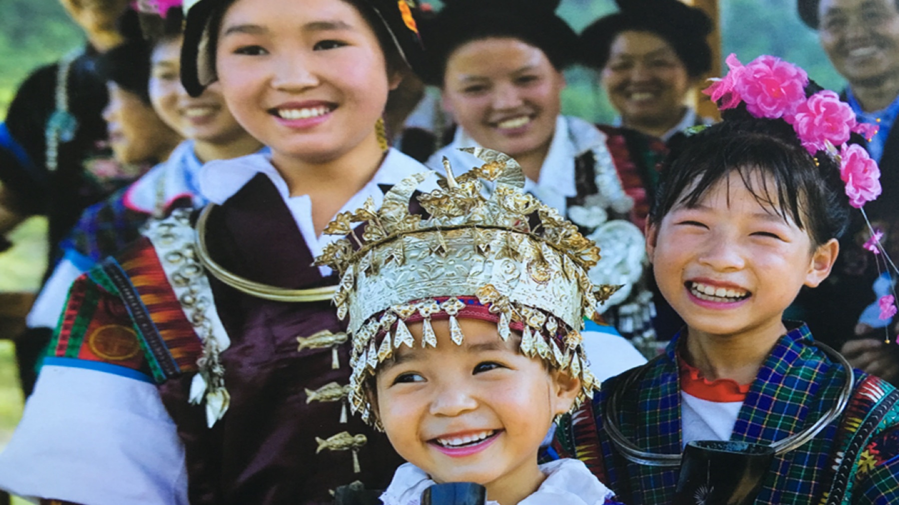 Three smiling children wear embroidered clothing, silver ornaments, and flowered headpieces.