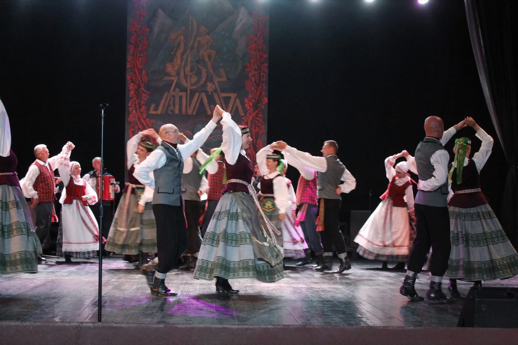 Adult folk dancers turning in pairs on a theatre stage while an accordion player performs.