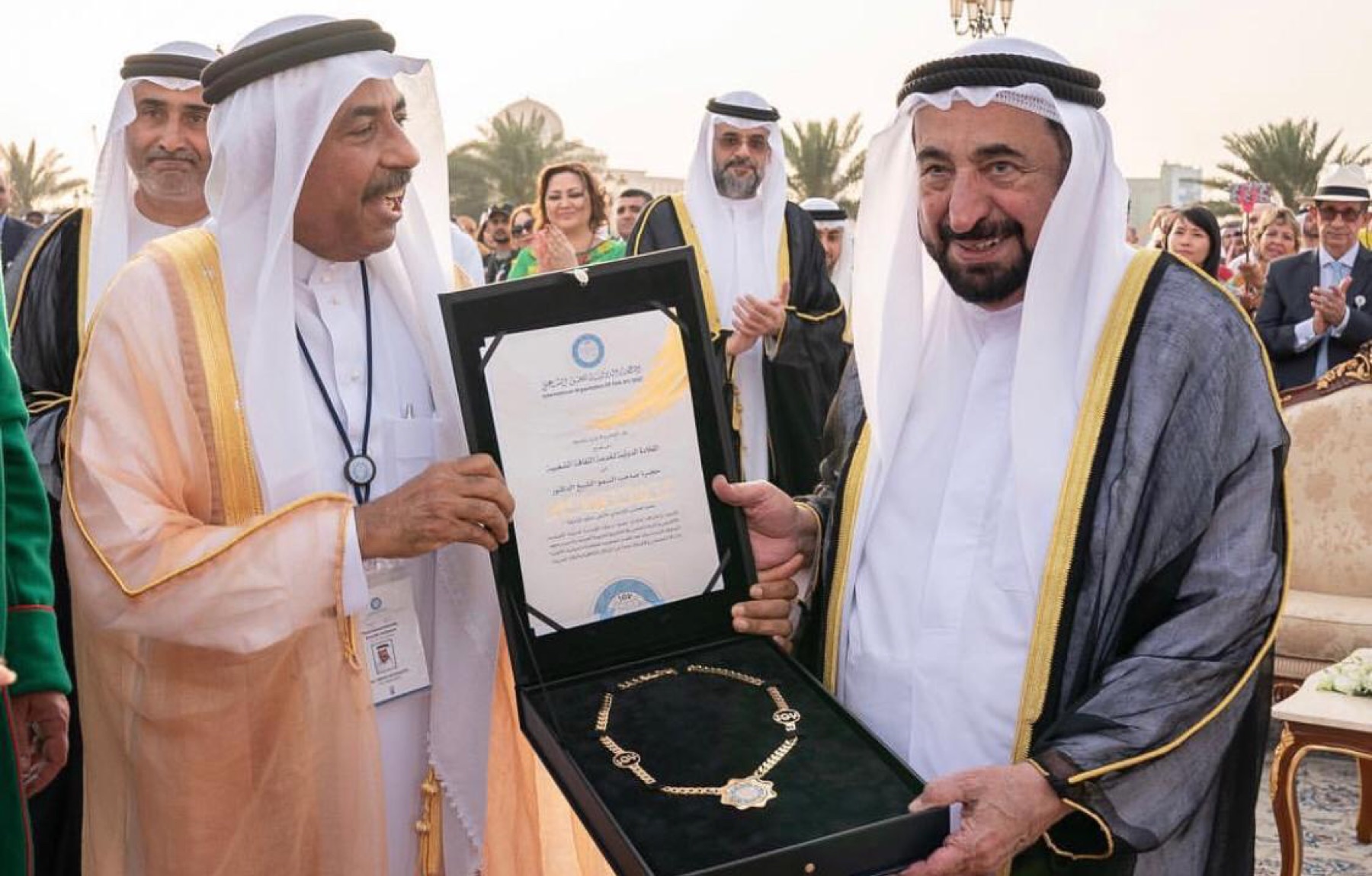Two men in formal Gulf dress display a framed recognition and a ceremonial gold chain.